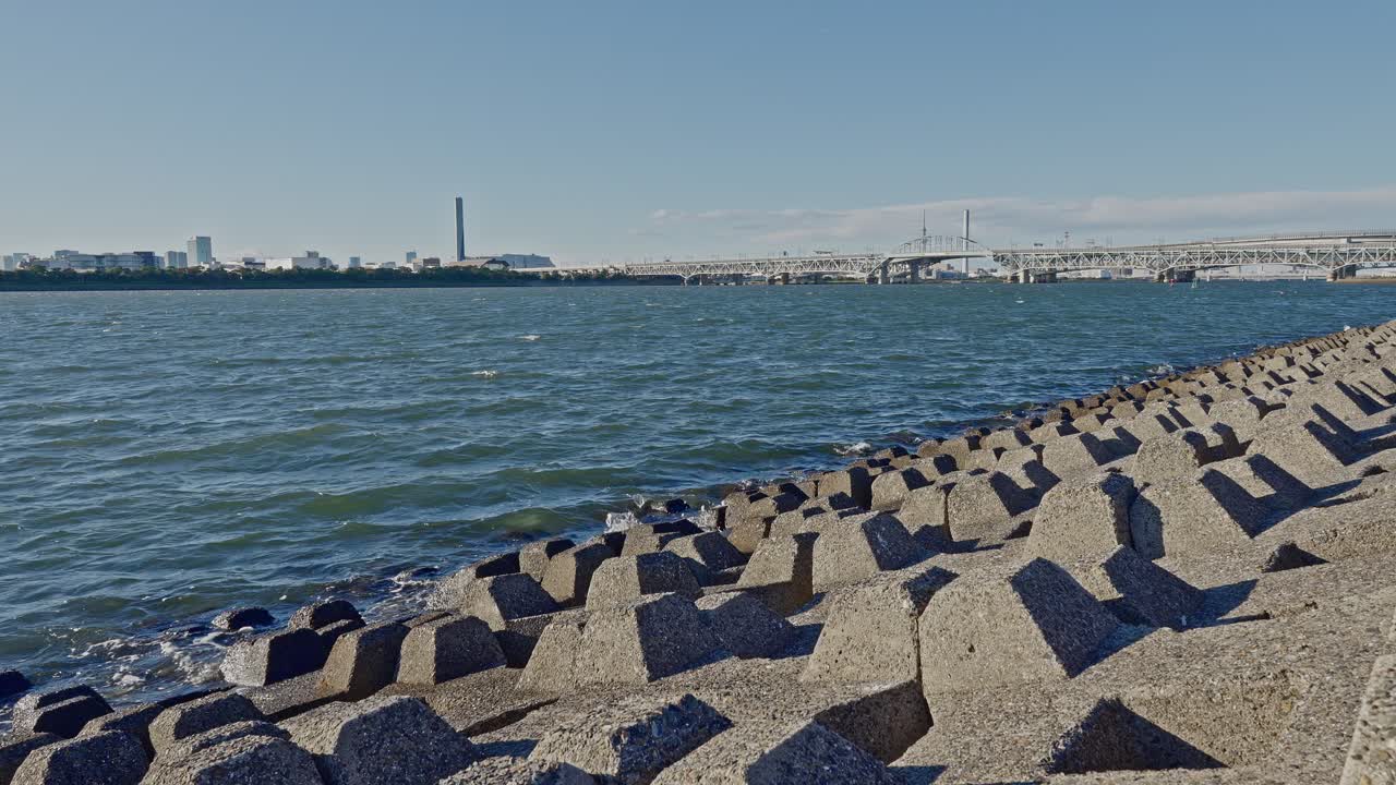 Daytime shot from behind large concrete tetrapods toward a wide bay with a modern bridge and distant city skyline