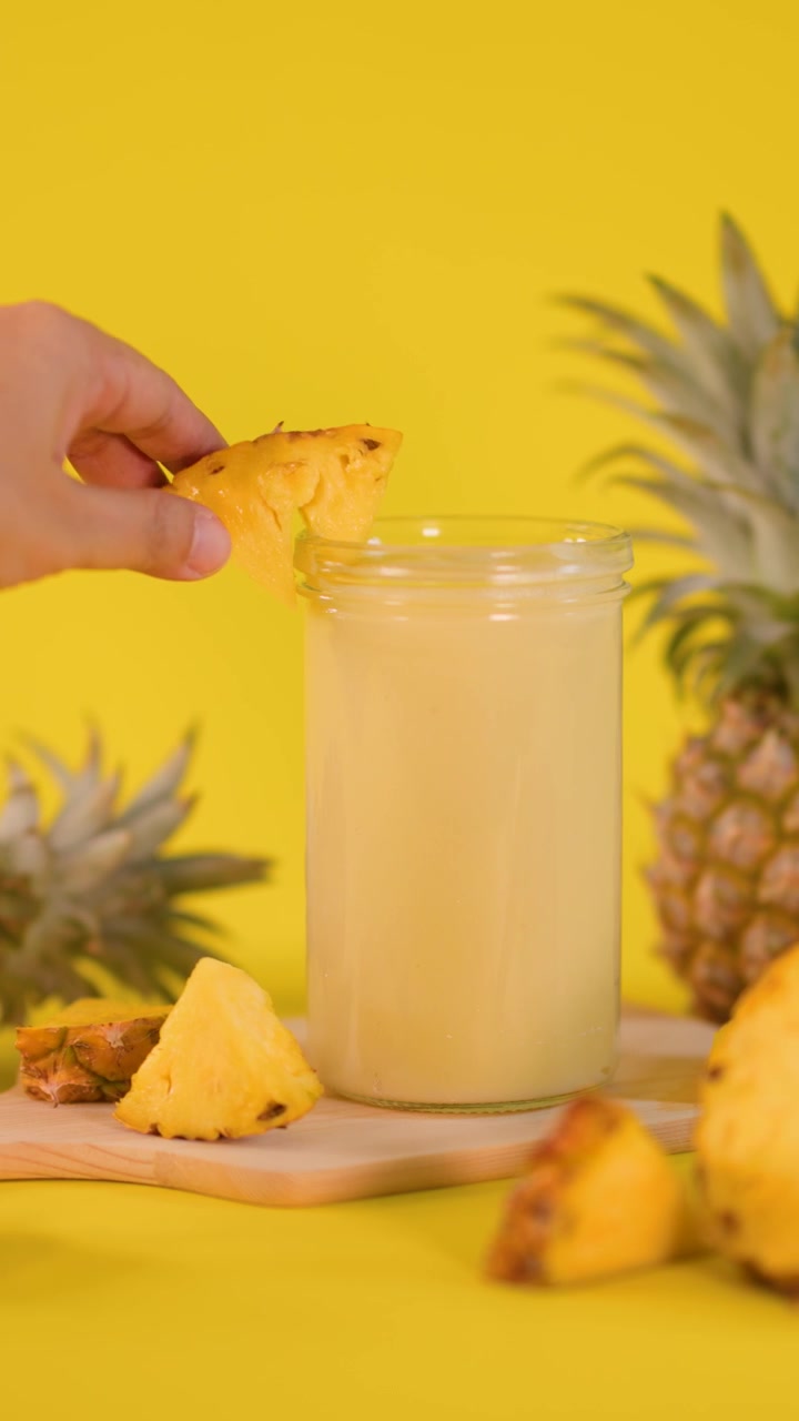 Una mano posiziona una fetta d'ananas su un frullato, circondato da frutta fresca, sfondo giallo brillante, inquadratura statica.