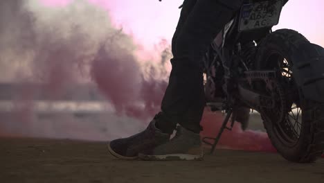 the biker is leaning on his bike while the engine is running and blowing out colored exhaust smoke, holi