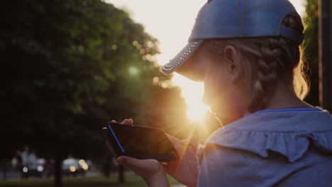 Girl-Sitting-In-The-Park-In-The-Evening-At-Sunset-Plays-On-The-Smartphone