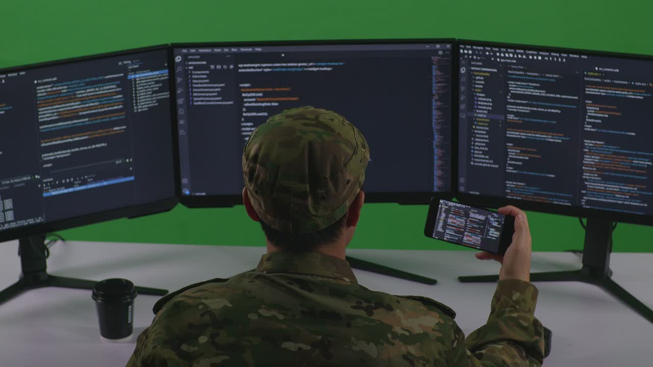 Back View Of Asian Man Soldier Developer Working With Mobile Phone While Write Code With Multiple Computer Screens In Green Screen Studio