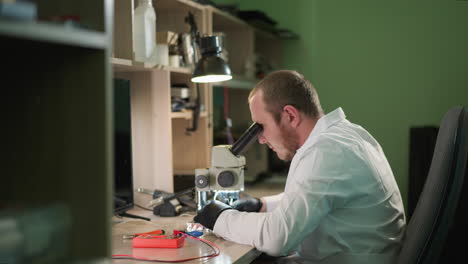 un técnico enfocado en una bata de laboratorio blanca examina algo bajo un microscopio en un entorno de laboratorio, el espacio de trabajo está equipado con instrumentos de trabajo, una lámpara de escritorio