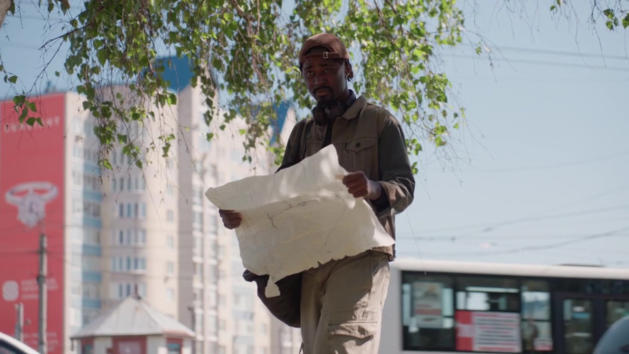 Close up of man examining large paper map on street with cars passing by and buildings in background. Man holds bag over shoulder while focusing on document under bright daylight. Urban