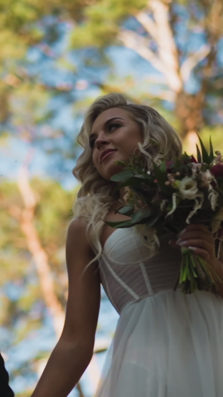 Happy new family of handsome man in suit and pretty woman bride with flowers bouquet walks joining hands against green park trees slow motion low angle shot