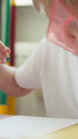 diligent toddler boy draws circle with plastic mould and pencil in notebook in classroom closeup. preschooler child learns simple shapes in nursery