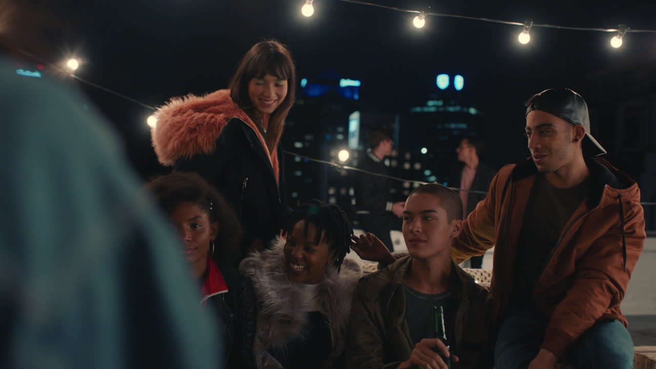 젊은 행복한 친구들이 밤에 옥상에서 사진을 찍고 파티를 즐기고 있습니다. 아프리카계 미국인 여성이 스마트폰을 사용하여 재회 모임을 촬영하고 소셜 미디어에서 공유하고 있습니다.