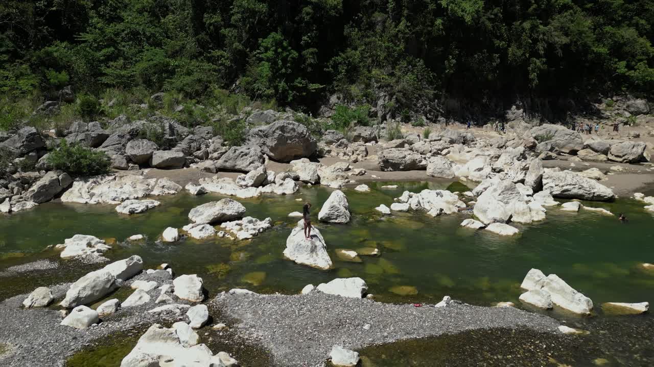 A woman balances on a rock at Tinipak River, striking a graceful pose before blowing a flying kiss as the drone pulls away—capturing both elegance and raw natural beauty