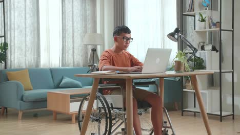 young asian boy sitting in a wheelchair using laptop computer while looking and smiling to camera at home