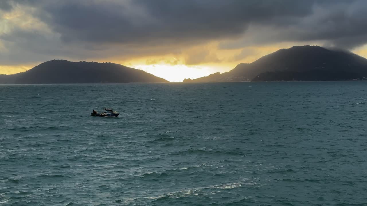 Fishing boat on choppy teal waters at sunset in the Gulf of La Spezia, Liguria, with dramatic clouds framing the channel between Palmaria and the Porto Venere headland