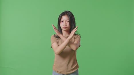 young asian woman with bruise on face looking into camera showing hands cross gesture to stop violence on green screen background in the studio