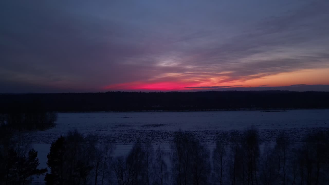En 4K-drone nærmer sig en tynd linje af ensomme træer på en snedækket eng i den dybe skumring. En sort skovsilhuet dominerer horisonten under en pink efterglød og en falmende blå himmel, lige efter solnedgang.