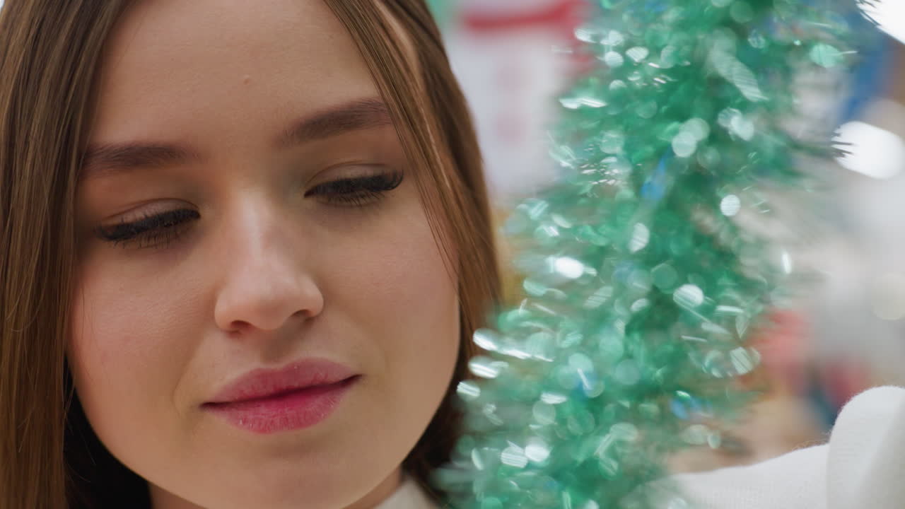 Close-up of woman with warm smile inspecting green shimmering tinsel, bokeh light effect adding festive ambiance in blurred background of decorative retail store