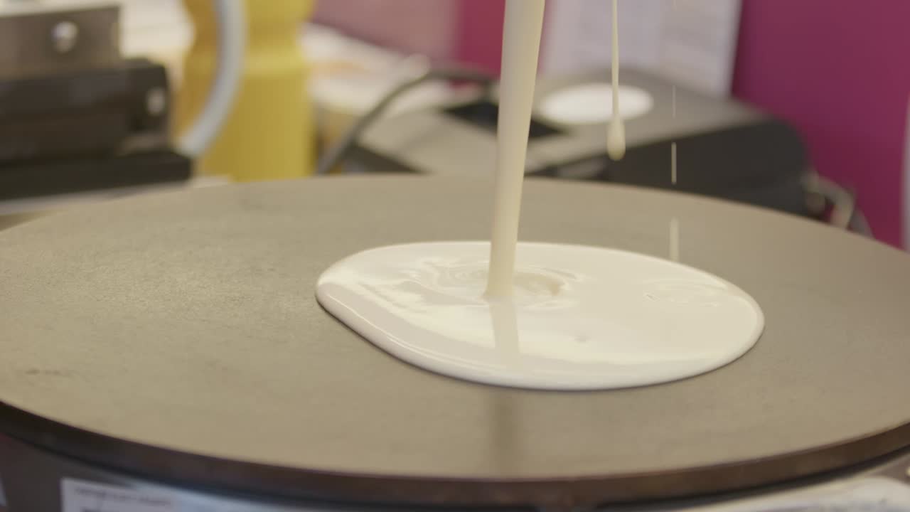 A close-up, static shot of thick, white crêpe batter being poured in a slow, steady stream onto the center of a hot, black round griddle. The liquid batter spreads slightly