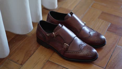 polished brown groom shoes with double buckle resting on parquet floor by curtain