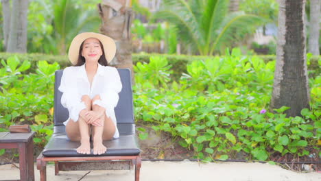 Mujer-Asiática-Sentada-En-Una-Tumbona-En-Un-Exótico-Salón-De-Playa-Con-Palmeras-En-El-Fondo,-Vestida-Con-Una-Blusa-Blanca-Y-Un-Sombrero-De-Paja---Espacio-Para-Copiar