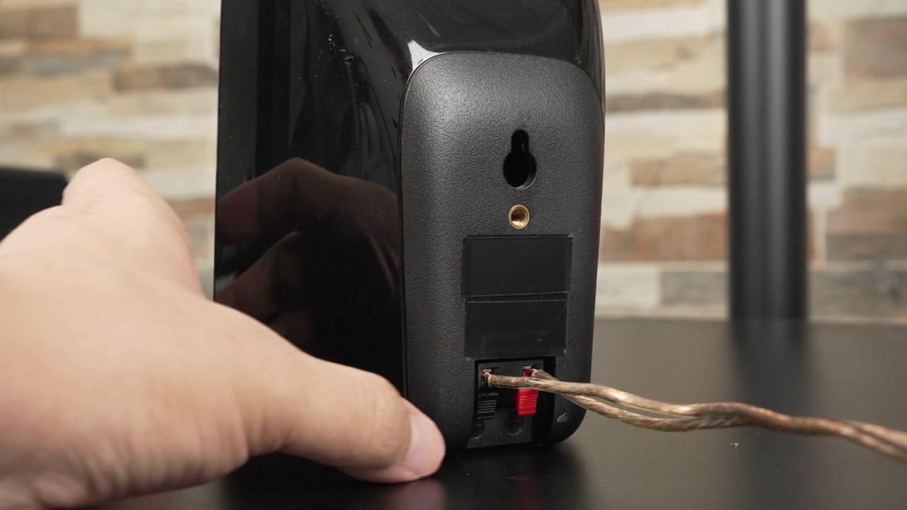 Hands attach bare speaker wire to the spring clip terminals on the back of a black bookshelf speaker on a desk, useful for tutorials, product demos, or home o guides.