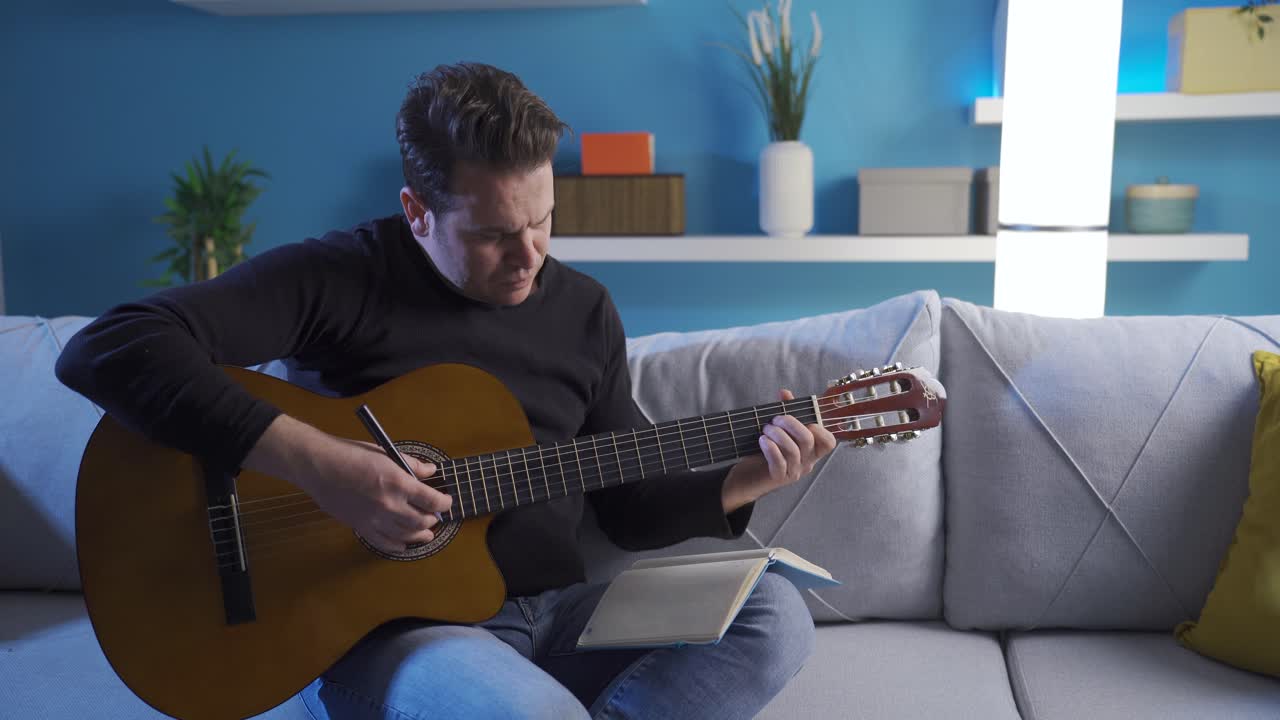 músico masculino sentado en el sofá en casa, tocando la guitarra y escribiendo letras en el cuaderno mientras compone una nueva canción.