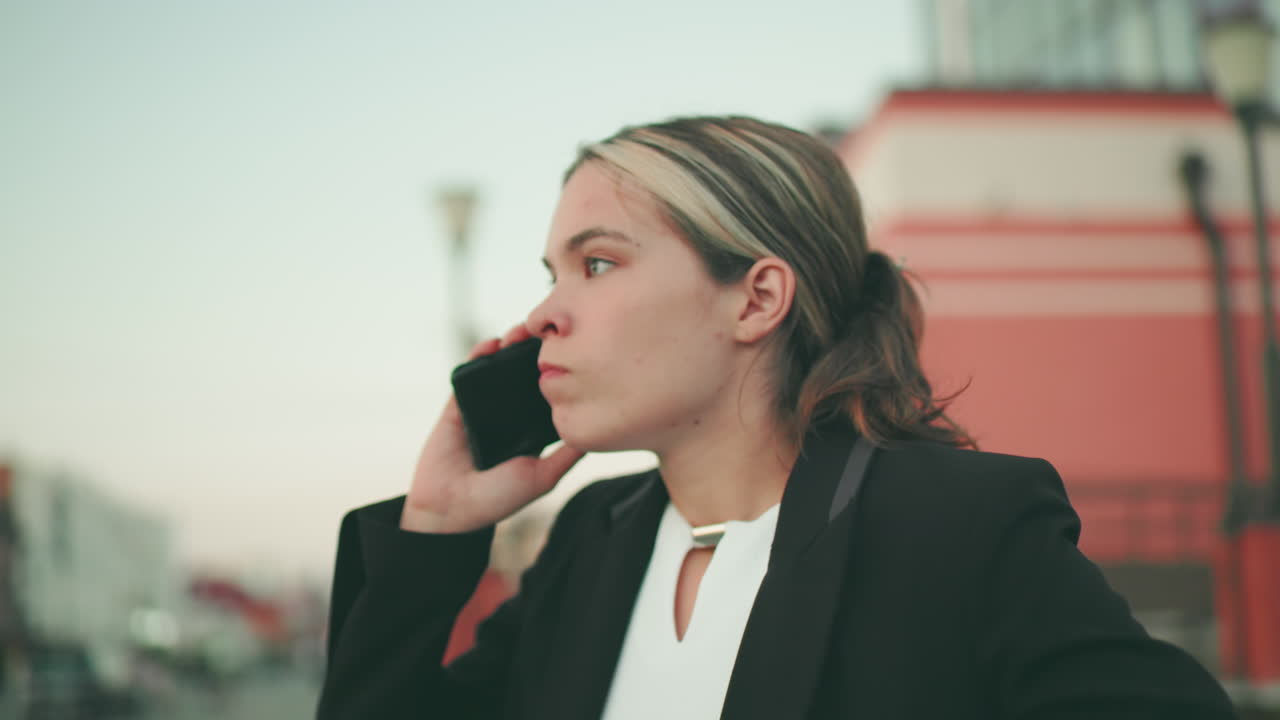 Furious lady in professional outfit appears visibly upset while on phone call in urban environment, people blur in distant background, displaying frustration