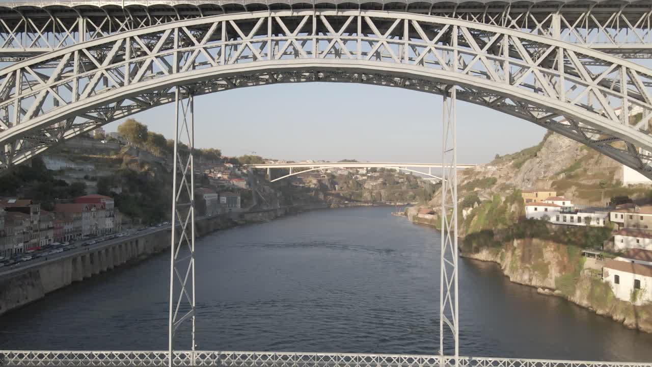 Aerial footage shows Dom Luis I Bridge spanning Douro River near Pinhao with metro train crossing upper level. Clear sky and distant hills form scenic backdrop to iconic steel arch structure