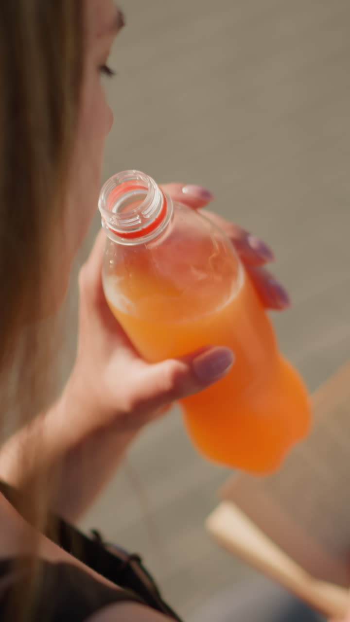 밖에서 앉아서 한 손으로 책을 들고 다른 손으로 병에서 주스를 마시고 있는 여성의 클로즈업, 책이 약간 흐릿하고, 그녀는 배경도 흐릿하게 읽고 있습니다.