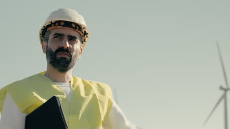 a focused engineer in a white helmet and reflective vest examines wind turbines in a field of clean energy generators, highlighting the ongoing effort to save our planet through renewable energy