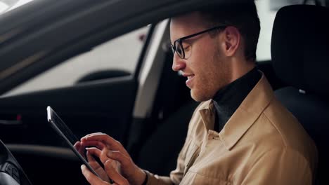 un joven elegante sentado en el auto detrás del volante pasa tiempo en la tableta