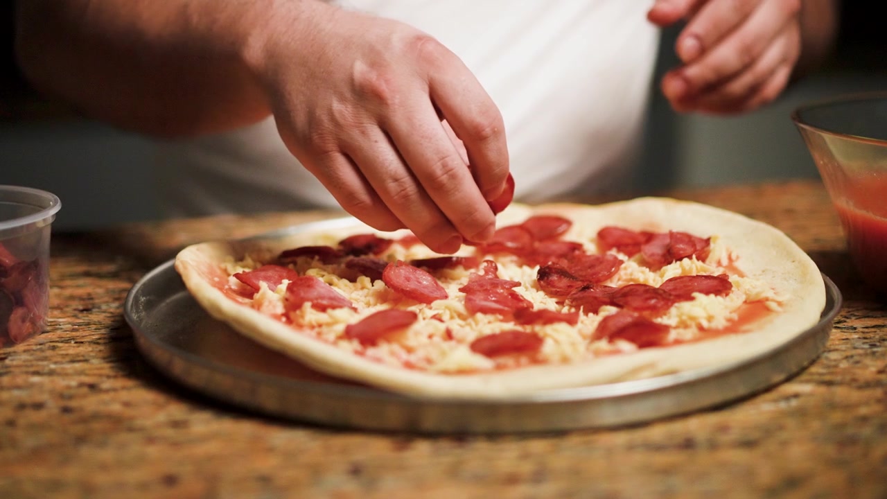 Preparazione artigianale della pizza con uno chef che aggiunge fette di pepperoni a una base di pizza appena stesa.