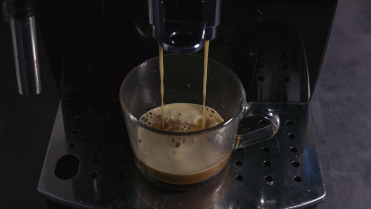 A glass cup sits beneath a coffee machine as two streams of espresso flow into it, creating a rich layer of crema on top. The close-up shot highlights the brewing process and the texture of the coffee