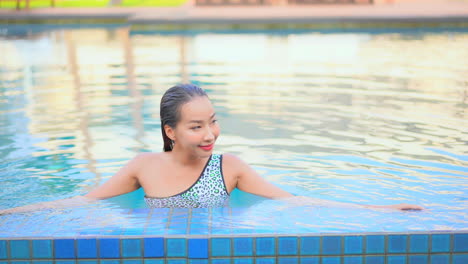 asian young woman leaning on the border inside swimming pool smiling, - front view daytime elevated view, template copy space
