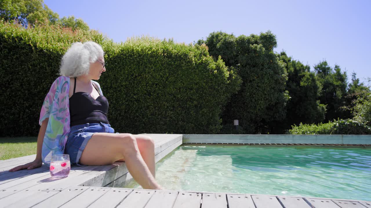 Senior woman arriving poolside dipping feet adjusting cover-up sipping cup and relaxing, copy space