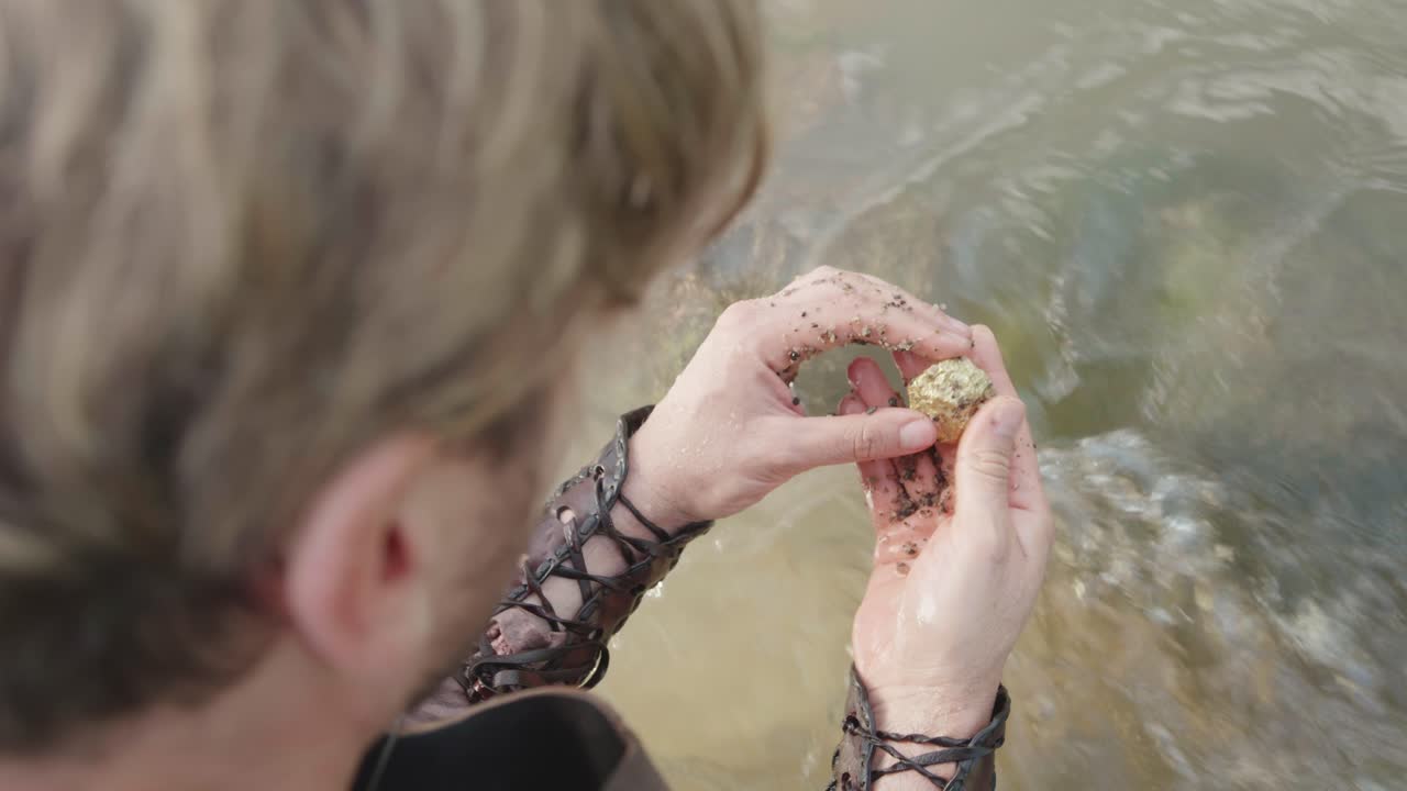 Over-the-shoulder shot of a historical prospector panning for gold. He sifts river sand in his hands and discovers a valuable, raw gold nugget. A concept of treasure hunting