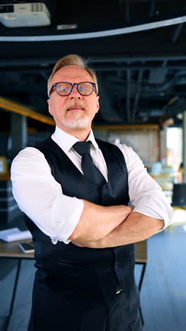 confident adult bearded businessman wearing white shirt and black vest stands in the office. man crossed hands on his chest thinking over something. vertical video.
