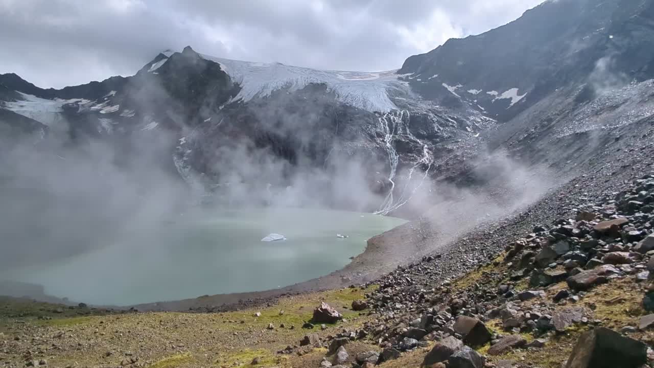 nubes y niebla rodando sobre un lago alpino remoto con un enorme glaciar por encima y la luz del sol rompiendo a través