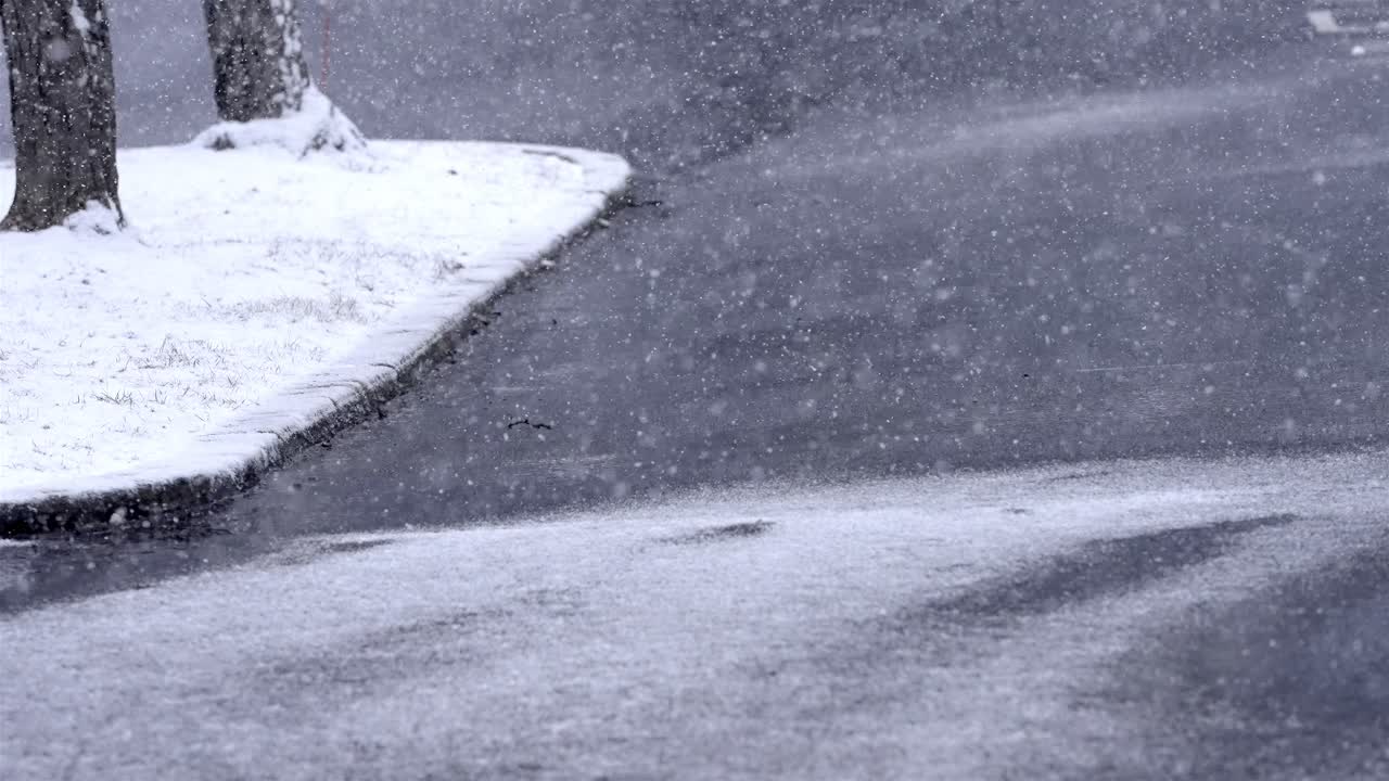 Slow motion snowfall on a street, road, asphalt on a winter morning, snowflakes falling cold weather, static shot, background, snow covered trees and grass