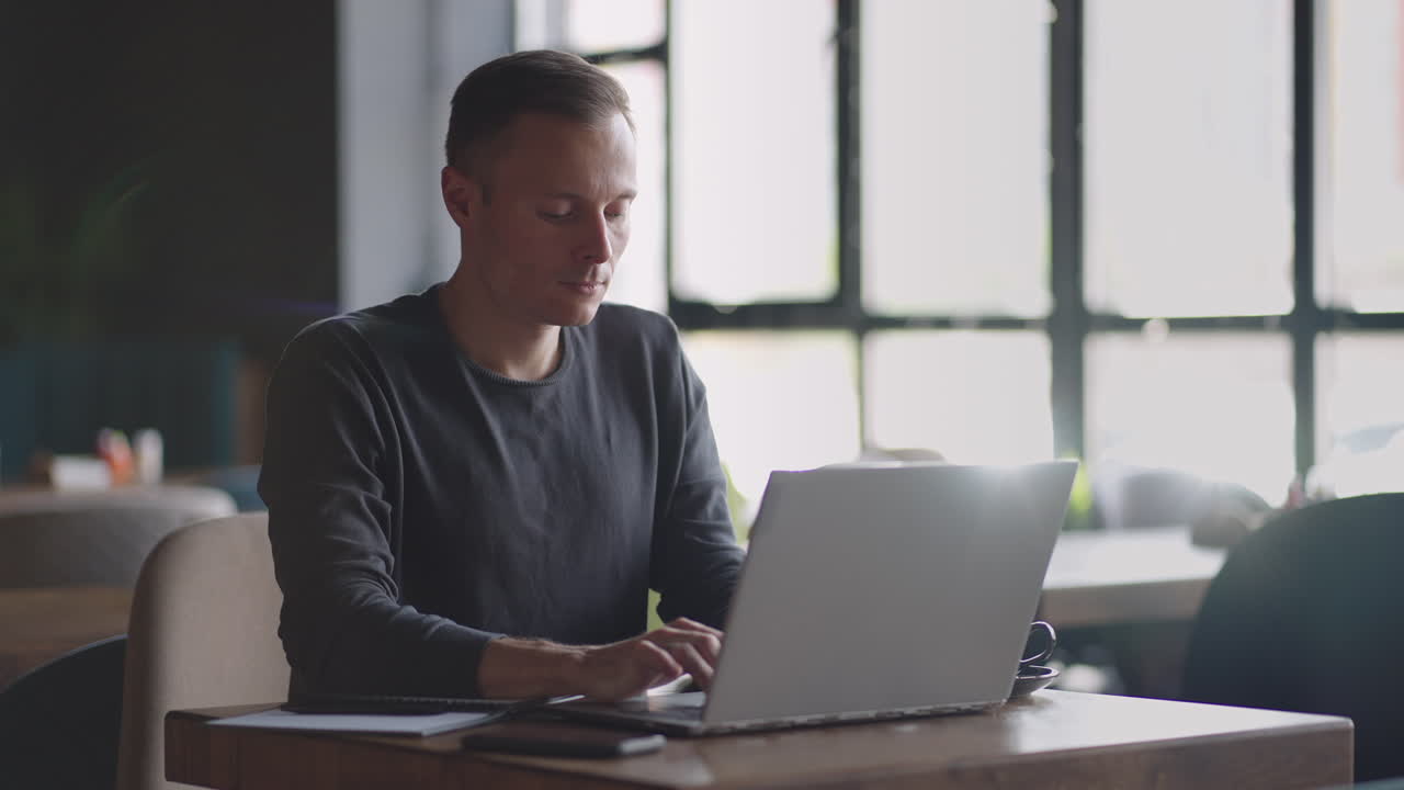 A young man in a shirt is sitting at a table with a laptop and typing on the keyboard. A student can study remotely. A businessman conducts his business remotely
