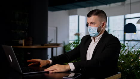 businessman in black suit works at his laptop wearing a mask. boss typing on his computer sitting at the table. dark blurred backdrop.