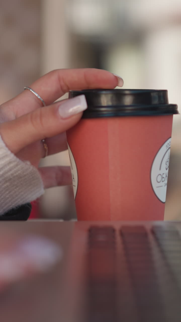 primer plano de la mano de la dama girando suavemente la taza de café cerca de la computadora portátil, centrándose en los dedos y anillos cuidados contra el teclado de la laptop, con el fondo borroso de la silla
