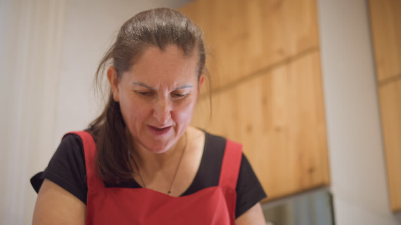 Donna bianca cuoca casalinga che prepara un pasto in una cucina accogliente, con un grembiule rosso, concentrata a mescolare e assaggiare, poi si apre in un sorriso gioioso. Mobili in legno e luce calda incorniciano un'intima scena domestica.