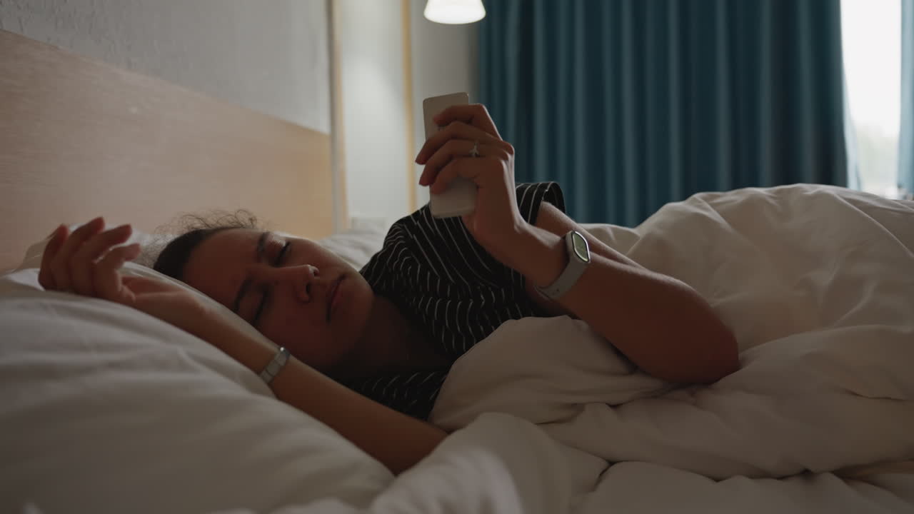 Kvinde hviler på hotelseng og tjekker smartphone, dæmpet sengelampe, blå gardiner, bløde hvide lagner, prikket kjole, armbånd, langsom opvågning, rejseopholdstemning, intimt morgenlys, afslappet alene.