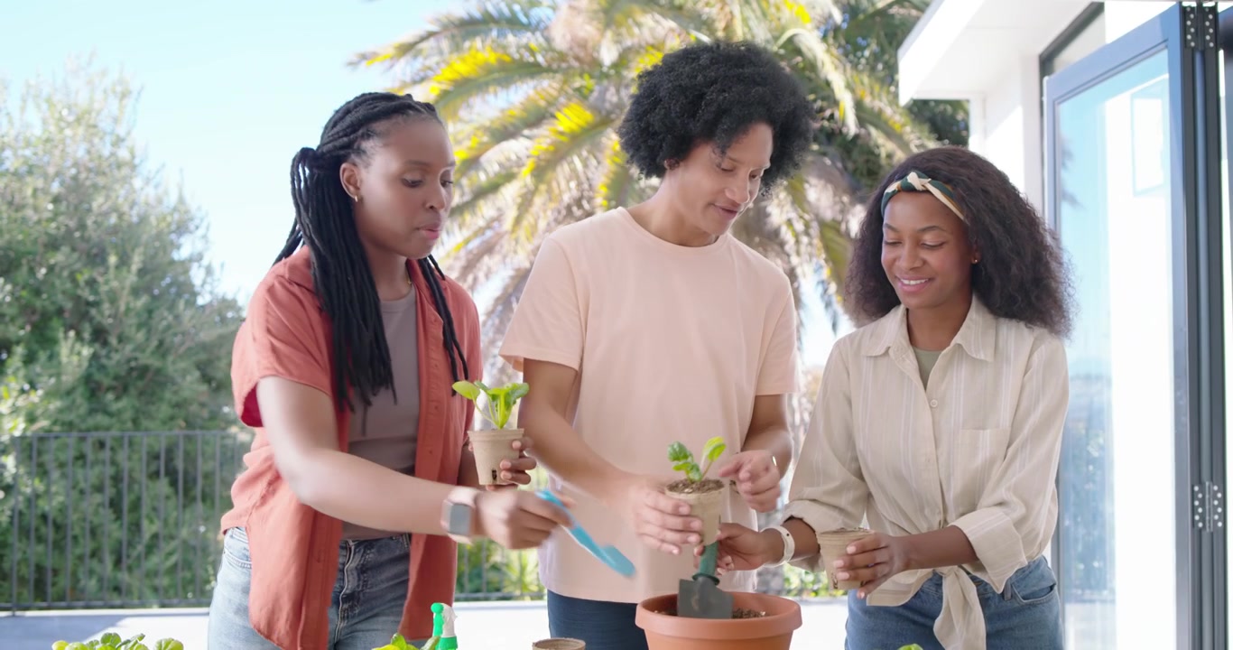 African American family handing tools, scooping soil and planting seedlings into clay pot on patio