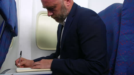 Hombre-Con-Barba-Escribiendo-En-Un-Cuaderno-Trabajando-En-Un-Avión-De-Pasajeros-Usando-Una-Mesa-De-Bandeja