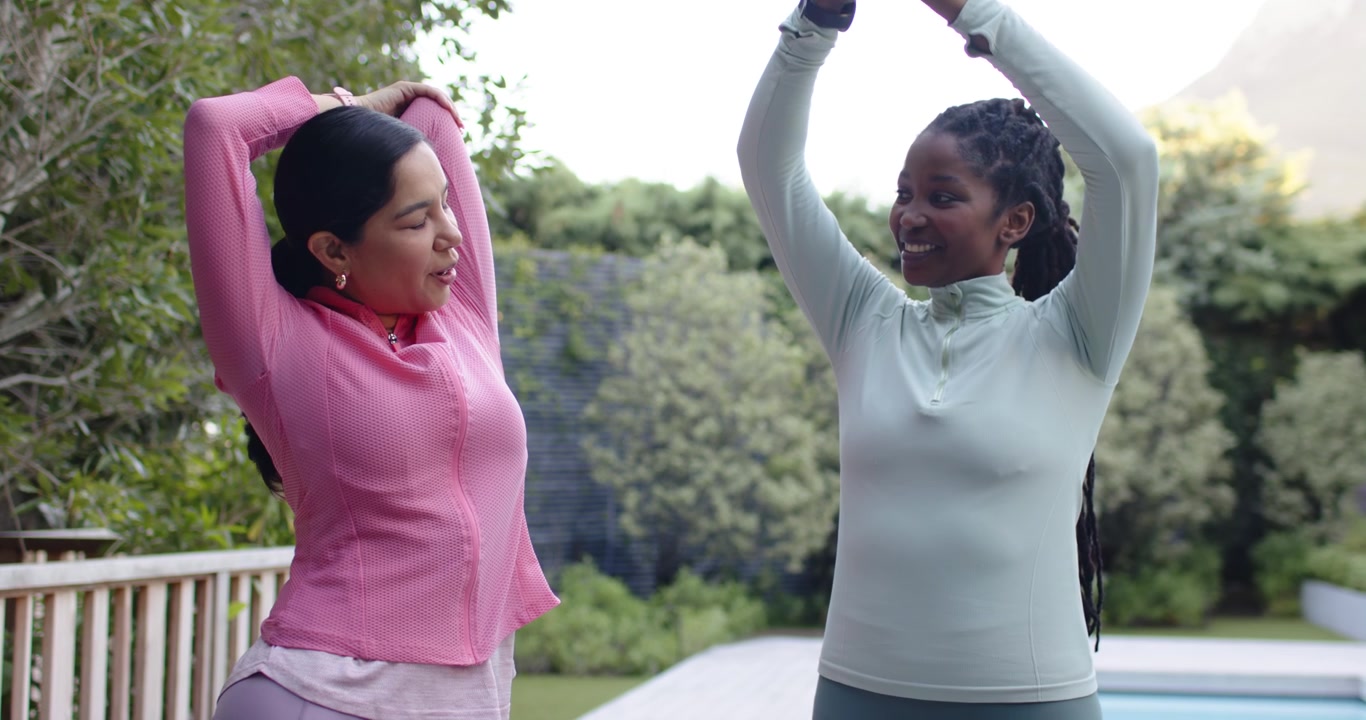 Amiche di diverse etnie fanno stretching sul bordo piscina, riscaldandosi per l'allenamento, in abbigliamento sportivo con tracker.