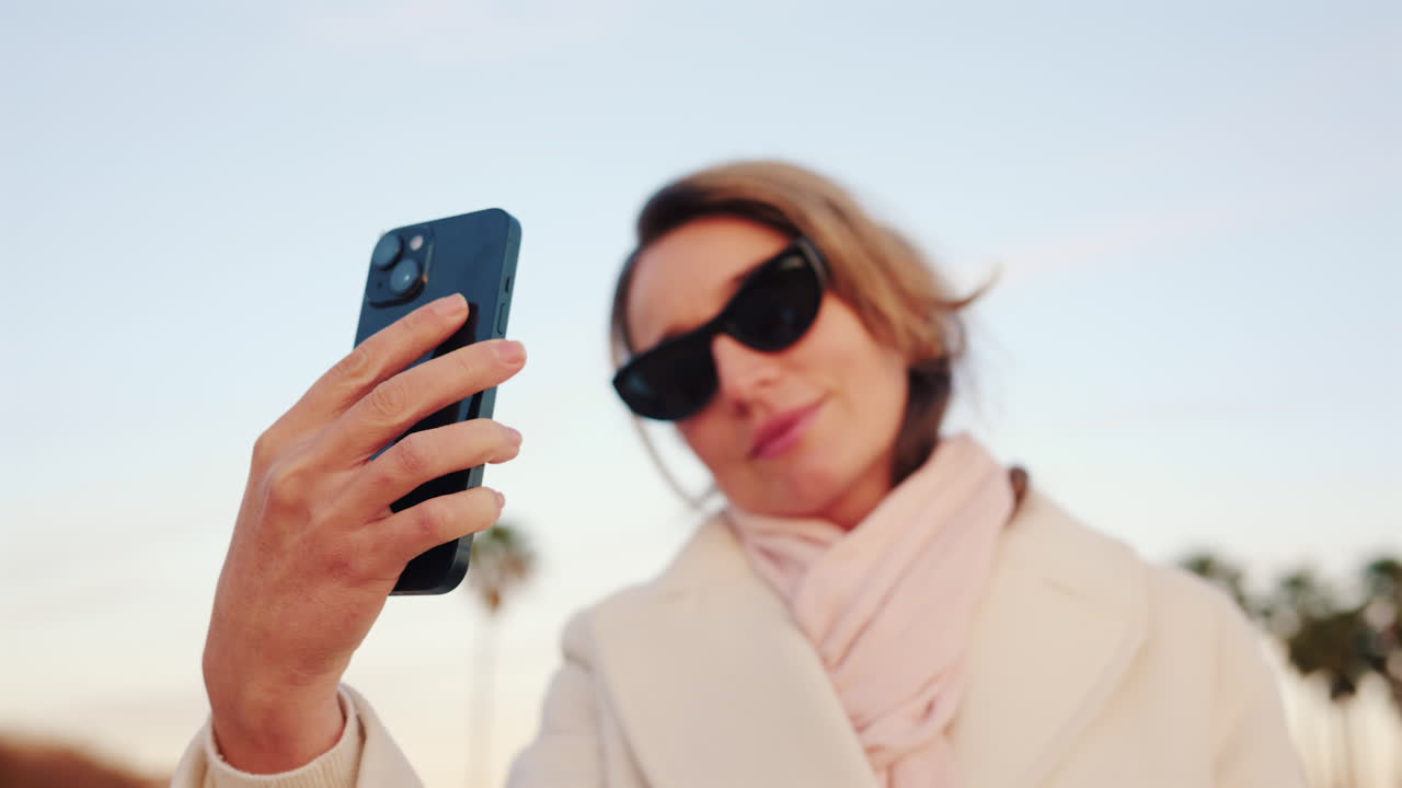 Confident woman in a white coat takes a selfie with her smartphone in a sunny outdoor area lined with palm trees