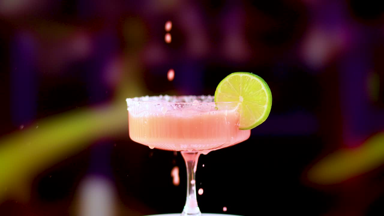 Uma margarita rosa é servida em um copo com uma rodela de limão, em um fundo colorido e desfocado.