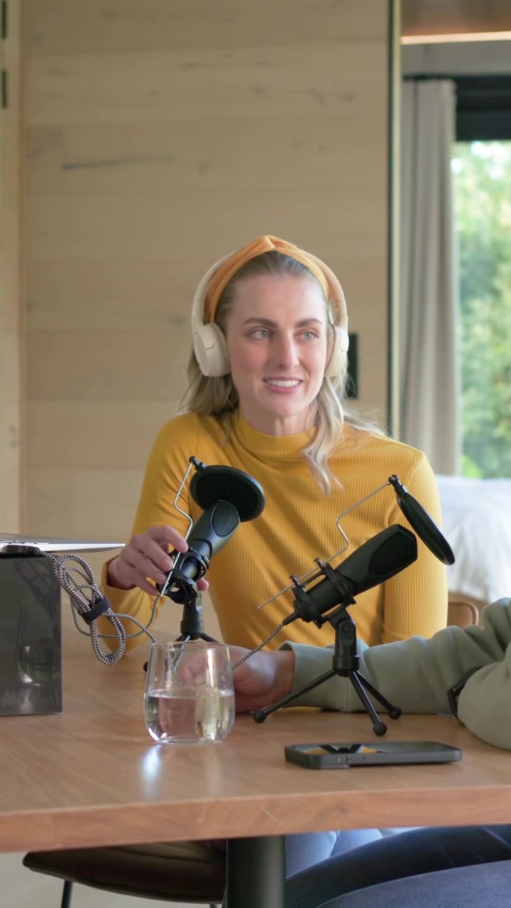 Video verticale: Donna con maglia color senape avvia un podcast, regola i telefoni al tavolo di legno e sistema il microfono.