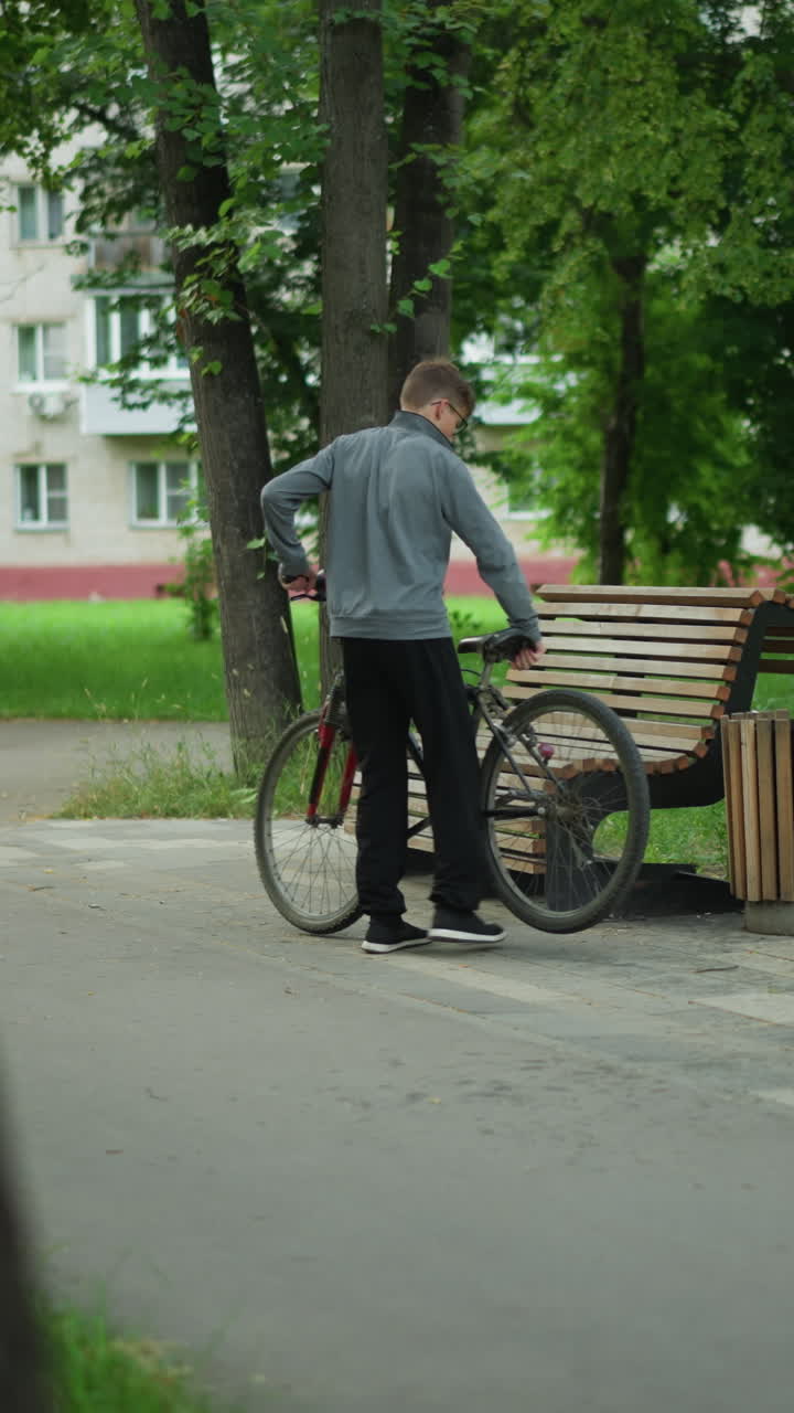 회색 을 입은 어린 소년은 조용한 나무로 둘러싸인 공원을 자전거로 타고, 나무 벤치에 멈추고, 자전거를 주차하고, 울창한 초록색으로 둘러싸여 도시 환경에서 자연을 즐기며 앉습니다.