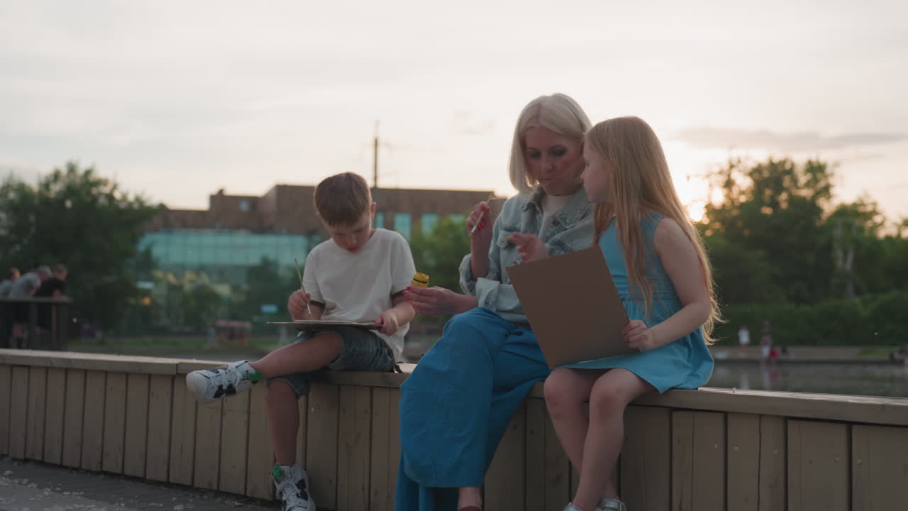young mom seated on wooden bridge between kids chatting painting with brush and jars on paper by pool under sunset glow, siblings holding boards while mother shares tips in outdoor art moment
