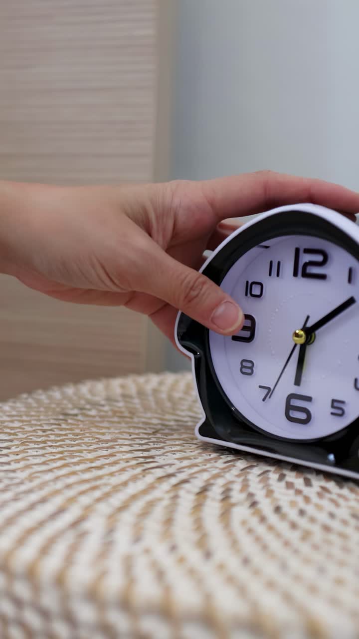 Una mano si allunga verso una sveglia in bianco e nero su un comodino intrecciato.