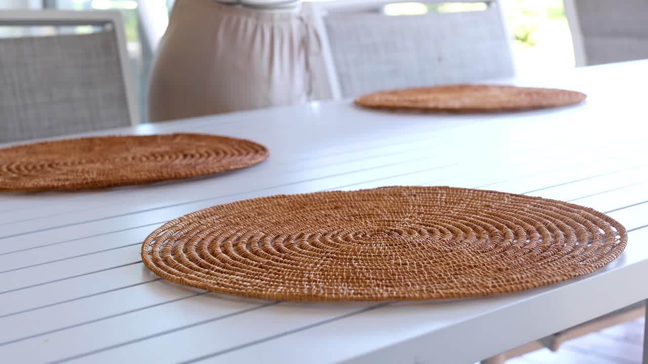 A person arranges woven placemats on a white table in a bright outdoor setting, creating a welcoming atmosphere