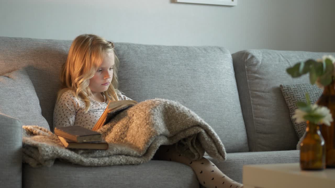 Ragazzina seduta sul divano che legge un libro, avvolta in una coperta.